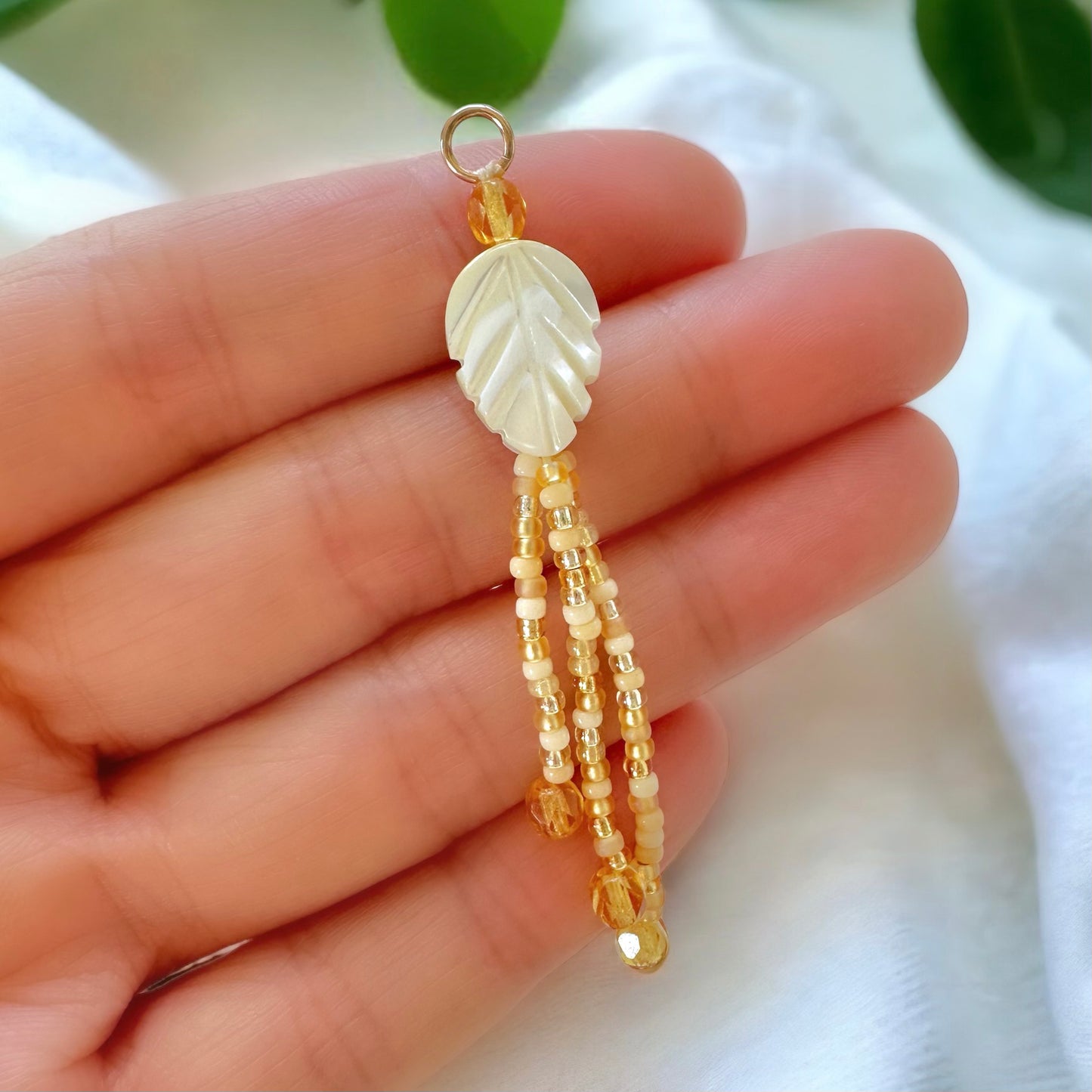 beaded fringe earring made with a mother of pearl leaf shaped bead and gold and cream miyuki 11/0 seed bead held in a woman's hand with a white cloth and green leaves in the background