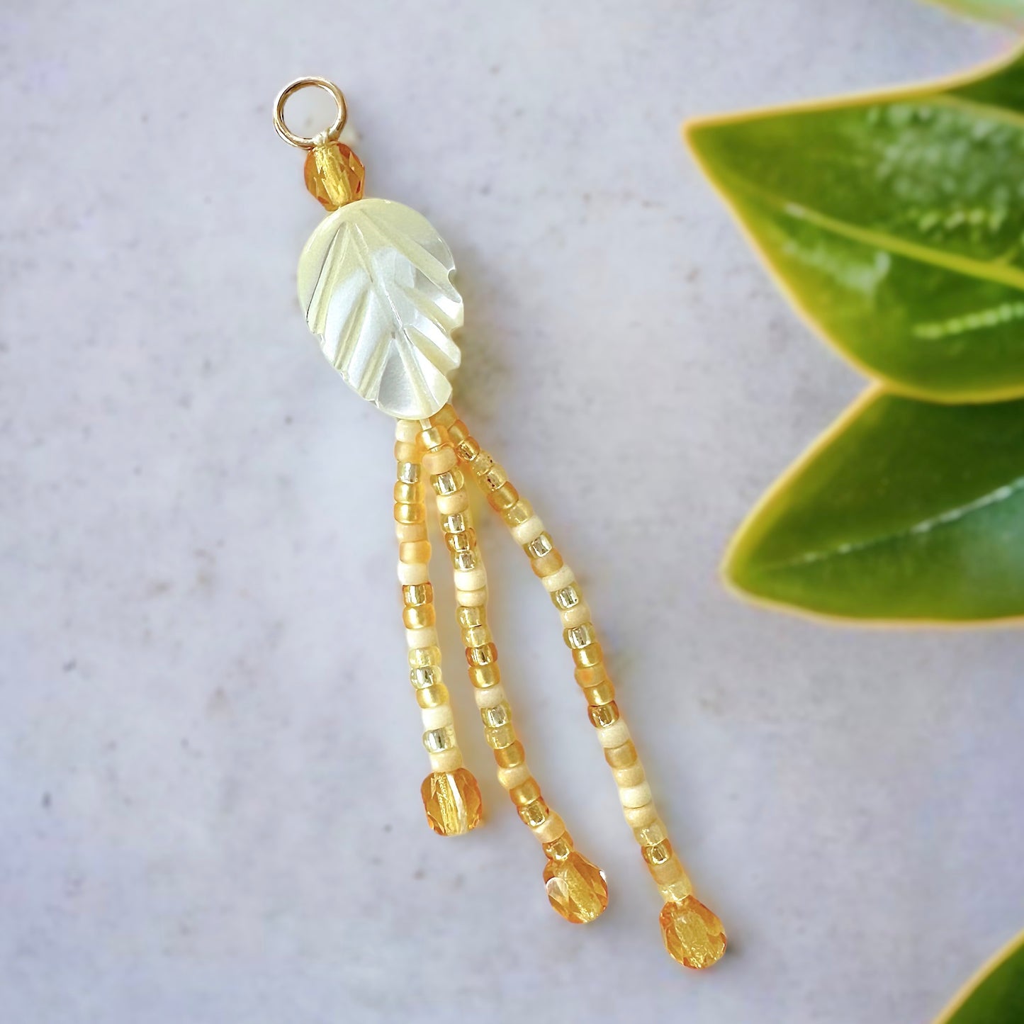 beaded fringe earring made with a mother of pearl leaf shaped bead and gold and cream miyuki 11/0 seed beads on a gray concrete surface with green leaves.